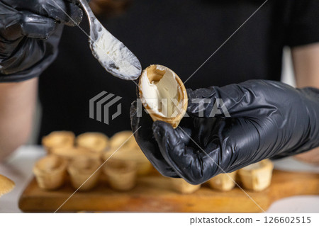 spreading creamy cheese into a crispy tartlet by a person wearing black gloves, preparing gourmet appetizers on a wooden board spreading creamy cheese into a crispy tartlet by a person wearing black gloves, preparing gourmet appetizers on a wooden board 126602515