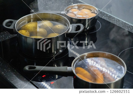 three pots with boiling potatoes, eggs, and carrots on a modern electric stovetop in a home kitchen, steam rising from the hot water during the cooking process three pots with boiling potatoes, eggs, and carrots on a modern electric stovetop in a home kitchen, steam rising from the hot water during the cooking process 126602551