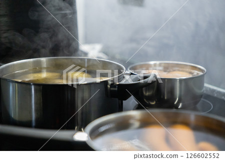 three pots with boiling potatoes, eggs, and carrots on a stovetop, steam rising as ingredients cook for a homemade meal preparation three pots with boiling potatoes, eggs, and carrots on a stovetop, steam rising as ingredients cook for a homemade meal preparation 126602552