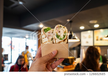 A Hand Holding a Takeaway Chicken Caesar Salad Wrap in Front of a Bustling Cafe Counter Showcases Fresh Grab and Go Dining Inside a Trendy London Eatery. A Hand Holding a Takeaway Chicken Caesar Salad Wrap in Front of a Bustling Cafe Counter Showcases Fresh Grab and Go Dining Inside a Trendy London Eatery. 126602685