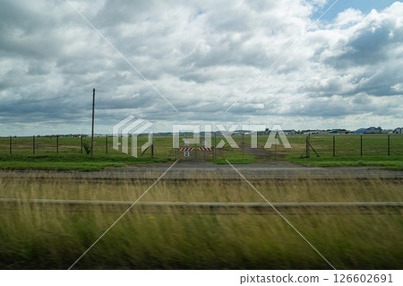 A Wide Grassy Field and Distant Airport Buildings Appear With Motion Blur Through a Tour Bus Window En Route to the Countryside Under a Cloudy Sky and Soft Natural Light. A Wide Grassy Field and Distant Airport Buildings Appear With Motion Blur Through a Tour Bus Window En Route to the Countryside Under a Cloudy Sky and Soft Natural Light. 126602691