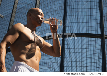 Athletes handsome sun-tanned shirtless man drinking water on outdoor sports ground. Concept water balance for hot season for active workout. Banner. Heat summer. Close up. Athletes handsome sun-tanned shirtless man drinking water on outdoor sports ground. Concept water balance for hot season for active workout. Banner. Heat summer. Close up. 126602936