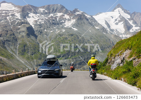 Cars and scooter drive Grossglockner High Alpine Road mountain green hills peaks blue sky background. Winding path through highland landscape offers scenic view and mountain driving experience 126603473