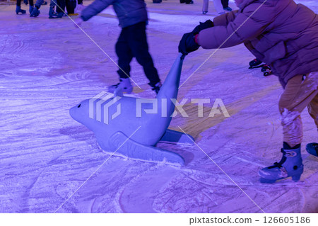 child using a seal-shaped skating aid on an ice rink during winter under colorful lights and snowy surface 126605186