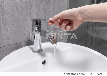 Hand turning on a modern chrome bathroom faucet with running water in a clean white sink Hand turning on a modern chrome bathroom faucet with running water in a clean white sink 126605260