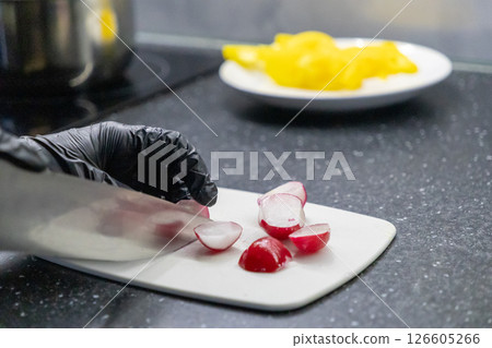 Slicing fresh radishes on a cutting board with a gloved hand and a sharp knife in a kitchen setting Slicing fresh radishes on a cutting board with a gloved hand and a sharp knife in a kitchen setting 126605266
