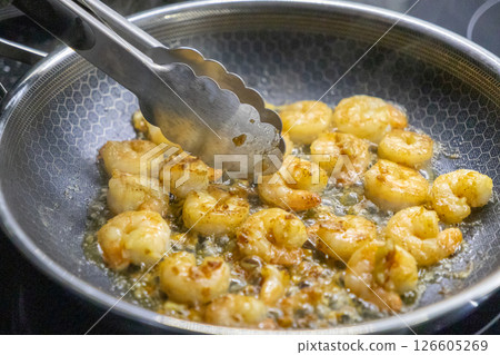 Juicy shrimp frying in a pan with tongs stirring, golden crust forming in sizzling oi Juicy shrimp frying in a pan with tongs stirring, golden crust forming in sizzling oi 126605269