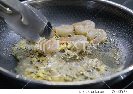 raw shrimp cooking in bubbling oil in frying pan with metal tongs holding one piece 126605283