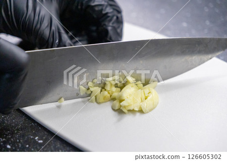 finely chopped garlic on white cutting board with sharp knife and gloved hand during food preparation finely chopped garlic on white cutting board with sharp knife and gloved hand during food preparation 126605302