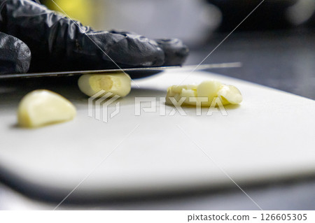 fresh garlic cloves being sliced on a white cutting board with sharp knife and gloved hand 126605305