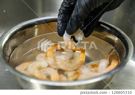 cleaned shrimp held by hand in black gloves over stainless steel bowl filled with water and shrimp in kitchen sink 126605310
