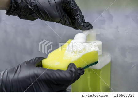 hand in black glove pressing soap dispenser onto yellow sponge with foam forming, cleaning preparation in kitchen setting hand in black glove pressing soap dispenser onto yellow sponge with foam forming, cleaning preparation in kitchen setting 126605325