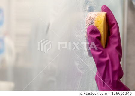 hand in pink rubber glove scrubbing glass surface with yellow sponge during bathroom cleaning for hygiene and sanitation 126605346