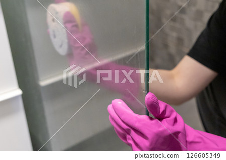 person in pink rubber gloves cleaning glass shower panel with yellow sponge, ensuring hygiene and clear surface in bathroom person in pink rubber gloves cleaning glass shower panel with yellow sponge, ensuring hygiene and clear surface in bathroom 126605349