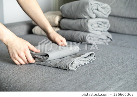 folding gray bedsheets by hand on textured fabric surface with neatly stacked linens in background for organized home aesthetic 126605362