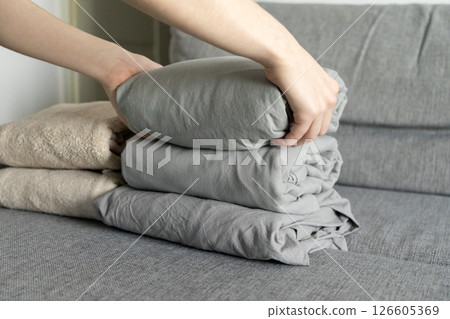 stacking gray linens on soft fabric sofa with hands carefully arranging folded bedding for tidy and organized home interior 126605369