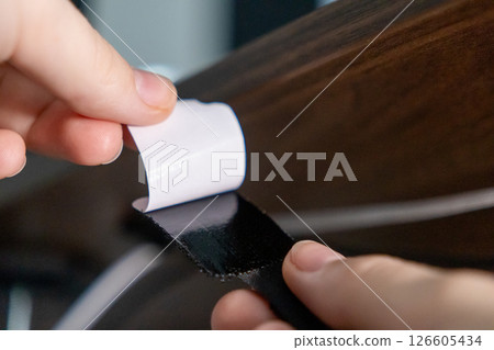 hand peeling adhesive backing from velcro strap to secure it under wooden desk for cable organization 126605434
