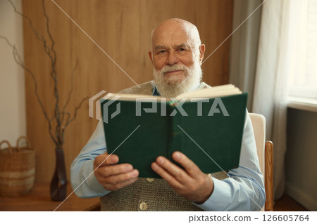 Serious retired senior man reading paper book sitting in cozy armchair at home 126605764