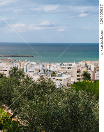 Olive trees frame picturesque view of coastal cityscape nestled against serene Mediterranean Sea on partly cloudy day in Crete, Greece. Olive grove overlooking coastal Cretan cityscape, Mediterranean 126605927