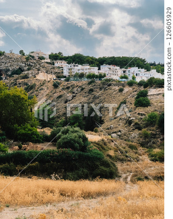 Dry grass and bushes growing on hillside with white houses on top of hill under cloudy sky in Rhodes, Greece, showing typical mediterranean landscape. Dry grasses, shrubs, Mediterranean hillside Dry grass and bushes growing on hillside with white houses on top of hill under cloudy sky in Rhodes, Greece, showing typical mediterranean landscape. Dry grasses, shrubs, Mediterranean hillside 126605929