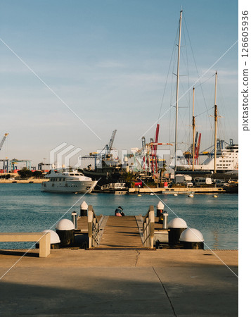 Concrete pier leading to luxury yachts and sailboats. Metal railed concrete pier extending toward luxury yachts, sailboats docked in harbor, industrial cranes framing nautical scene beneath azure sky Concrete pier leading to luxury yachts and sailboats. Metal railed concrete pier extending toward luxury yachts, sailboats docked in harbor, industrial cranes framing nautical scene beneath azure sky 126605936