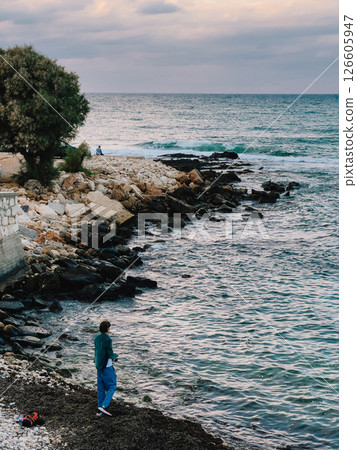 Young man standing on rocky shore, gazing at tranquil Mediterranean waters of Monopoli, Italy, as sun sets and casts warm glow over coastline. Young male standing on rocky coastline, rocky shore 126605947