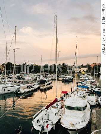Sailboats and yachts are moored in tranquil marina as sun sets, casting warm golden glow on water and creating picturesque and serene scene of nautical beauty. Sailboats rest in marina during sunset 126605950