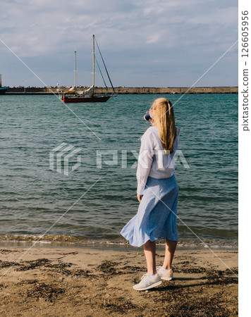 Blonde woman sipping coffee on beach, enjoying view of red sailboat gliding away on sunny day, framed by backdrop of fluffy clouds and endless blue sky. Blonde woman drinks coffee, red sailboat, ocean Blonde woman sipping coffee on beach, enjoying view of red sailboat gliding away on sunny day, framed by backdrop of fluffy clouds and endless blue sky. Blonde woman drinks coffee, red sailboat, ocean 126605956