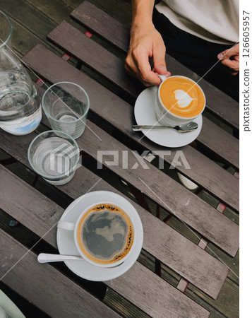 Cappuccino garnished with intricate latte art next to freshly brewed filter coffee on rustic wooden table surrounded by empty glasses and bottle of water. Latte art cappuccino, filter coffee, cafe Cappuccino garnished with intricate latte art next to freshly brewed filter coffee on rustic wooden table surrounded by empty glasses and bottle of water. Latte art cappuccino, filter coffee, cafe 126605957