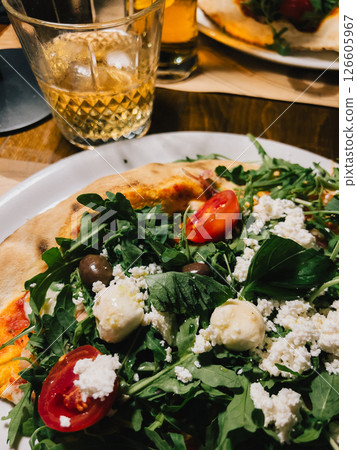 Close up of pizza topped with fresh arugula, juicy tomatoes, salty olives, crumbled feta, and creamy mozzarella on white plate, paired with refreshing alcoholic beverages. Beer, rustic white plate Close up of pizza topped with fresh arugula, juicy tomatoes, salty olives, crumbled feta, and creamy mozzarella on white plate, paired with refreshing alcoholic beverages. Beer, rustic white plate 126605967