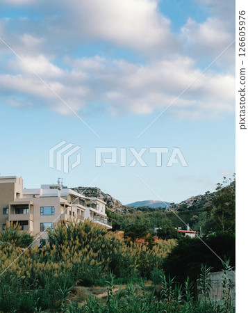 Lush green vegetation partially obscures modern hotel against rolling hills under dynamic, cloud filled sky, creating tranquil and inviting scene. Verdant landscape, undulating terrain, cloudscape 126605976