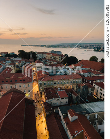 Sunset illuminating Zadar old town, revealing cobblestone streets bustling with tourists and Sea coastline stretching beyond historic urban landscape. Aerial view old town with illuminated street 126605983