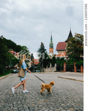 Young woman with long hair, wearing light blue summer dress and carrying reusable shopping bag, walking dog on cobblestone street in European city. eco friendly shopping bag. Young woman walking Young woman with long hair, wearing light blue summer dress and carrying reusable shopping bag, walking dog on cobblestone street in European city. eco friendly shopping bag. Young woman walking 126605986