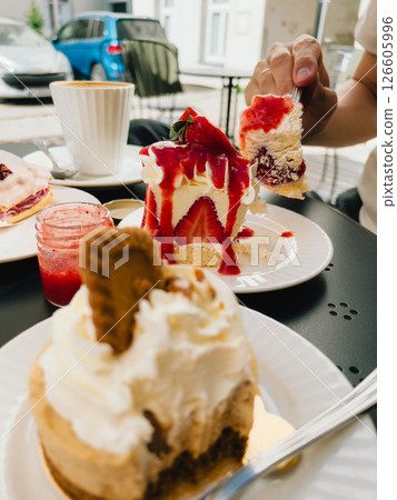 Hand holding fork with piece of delicious strawberry cake covered in whipped cream and strawberry sauce, another cake and cup of coffee on table in outdoor cafe. Silverware with creamy strawberry cake 126605996