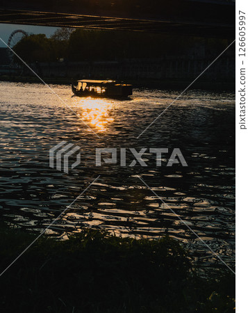 Sightseeing boat transporting tourists on river during golden sunset, with Ferris wheel and bridge in background, creating romantic atmosphere. Golden sunset, Ferris wheel, historic bridge, cityscape 126605997