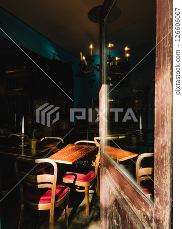 Sunlight streaming through weathered wooden door illuminates wooden tables and red upholstered chairs in cozy, vintage style restaurant, creating warm ambiance. Red chairs inside vintage restaurant 126606007