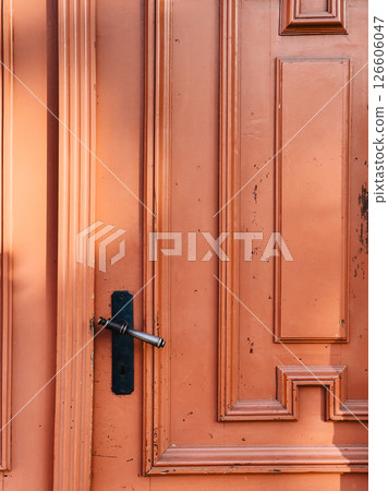 Sunlight illuminating old, weathered orange wooden door featuring classic metal handle and keyhole, showcasing texture and craftsmanship of a bygone era. Warm sunlight, weathered orange wooden door 126606047