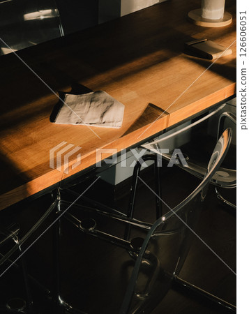 Sunlight illuminates a folded cloth tissue resting on a modern wooden table, with transparent chairs beneath, creating a scene of preparedness and adaptation in a contemporary workspace Sunlight illuminates a folded cloth tissue resting on a modern wooden table, with transparent chairs beneath, creating a scene of preparedness and adaptation in a contemporary workspace 126606051