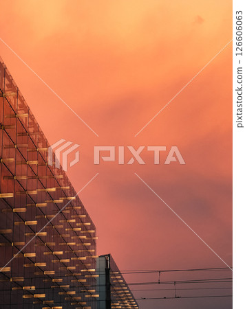 Glass facade of contemporary building reflecting warm orange hues of sunset sky, creating striking and vibrant architectural scene in urban cityscape. Glass facade reflecting warm orange sunset hues 126606063