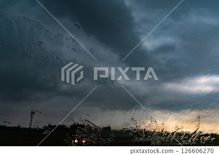 Rain shower on car windshield or car window and blurry road in background. Driving in rainy season. Rain drops on car mirror. Storm clouds, tornado, safety. Rain shower on car windshield or car window and blurry road in background. Driving in rainy season. Rain drops on car mirror. Storm clouds, tornado, safety. 126606270