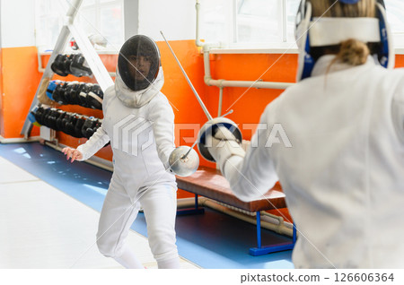 Female fencer facing opponent in close-up indoor duel. Female fencer facing opponent in close-up indoor duel. 126606364