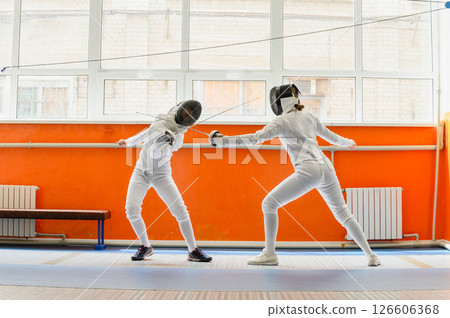 Female fencer striking opponent in bright indoor fencing hall. Female fencer striking opponent in bright indoor fencing hall. 126606368