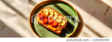 A vibrant sandwich features layers of strawberries, cucumbers, and nuts on toasted bread. This delightful treat is placed on a stylish plate in a sunlit kitchen setting, banner 126606564