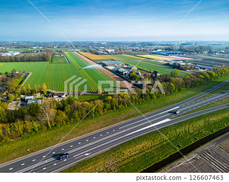 Aerial panoramic view of the Dutch countryside featuring farmland, green fields, forests, and a highway stretching through the landscape toward a distant town. 126607463