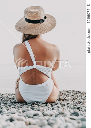 A woman is sitting on a beach wearing a straw hat and a white bikini. The beach is rocky and the sky is blue. A woman is sitting on a beach wearing a straw hat and a white bikini. The beach is rocky and the sky is blue. 126607548