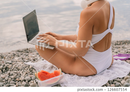 A woman is sitting on the beach with a laptop open in front of her. She is wearing a white bikini and has a pink headband on. Concept of relaxation and leisure. A woman is sitting on the beach with a laptop open in front of her. She is wearing a white bikini and has a pink headband on. Concept of relaxation and leisure. 126607554