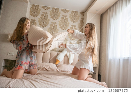 Pillow Fight Bedroom Fun: Two women playfully fight pillows on a bed in a modern bedroom, enjoying leisure time. 126607575