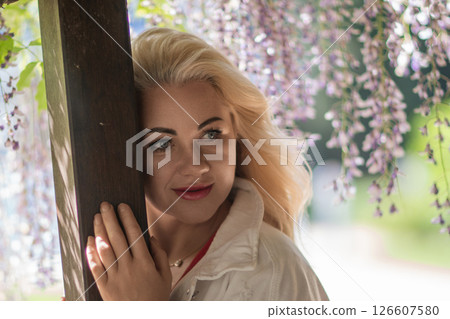 Woman wisteria portrait: Blonde woman leans on post in blooming wisteria garden during daytime. 126607580