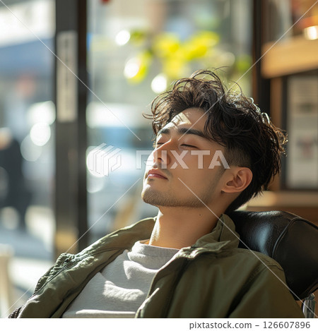 In a modern barber shop, an Asian man sits in profile, relaxed, as a barber styles his hair. Warm natural light highlights the scene, creating a serene atmosphere during the daytime 126607896