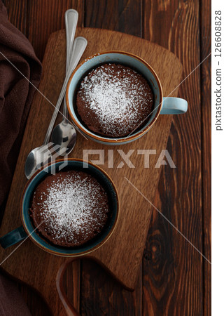 Two chocolate mug cake under sugar icing on dark wooden table. Easy homemade dessert. Mugcake from microwave. Top view Two chocolate mug cake under sugar icing on dark wooden table. Easy homemade dessert. Mugcake from microwave. Top view 126608828